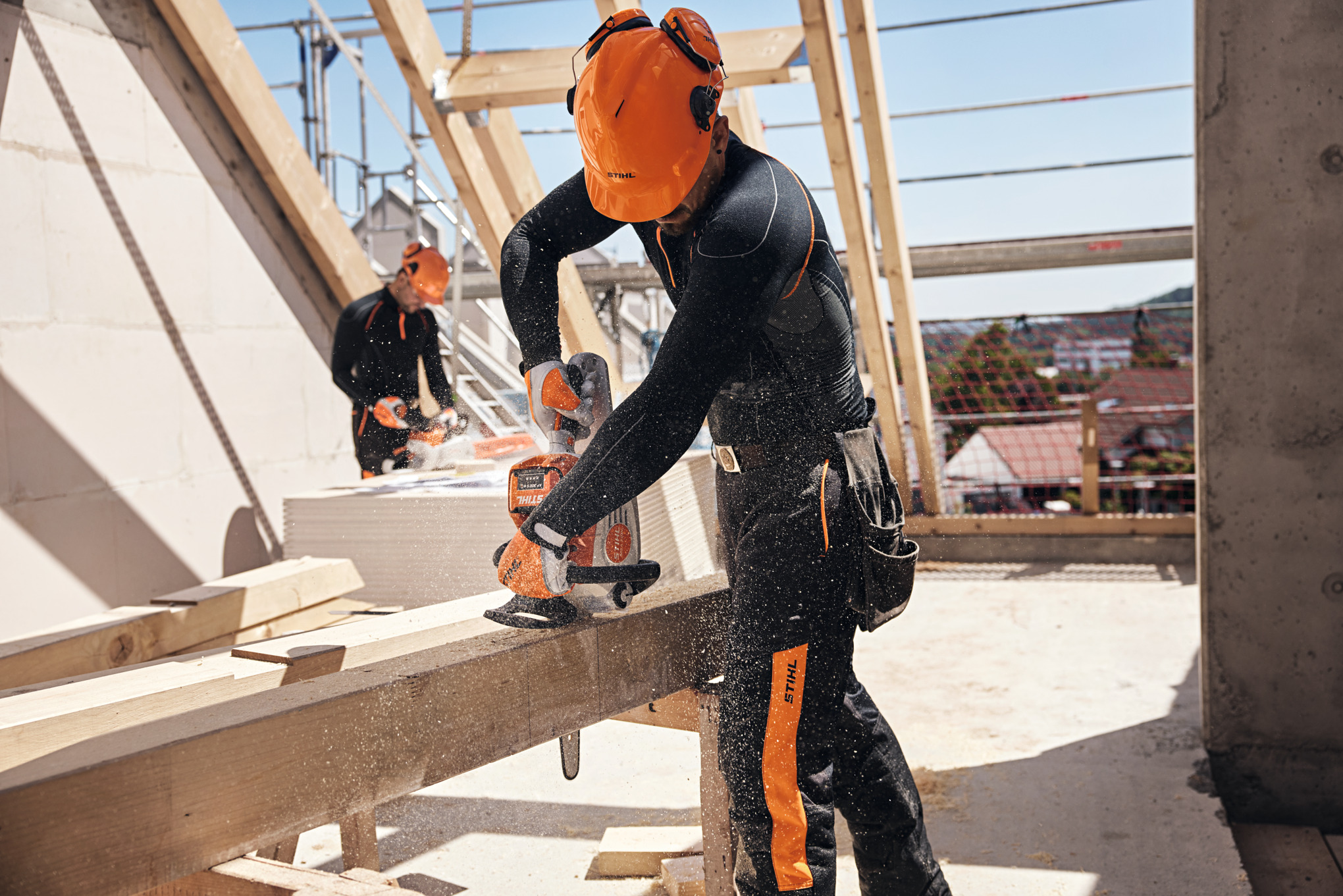 Ein STIHL Profi in einem Dachstuhl zersägt mit einer Akku-Motorsäge einen Holzbalken, im Hintergrund hantiert ein weiterer Arbeiter mit einer Akku-Motorsäge